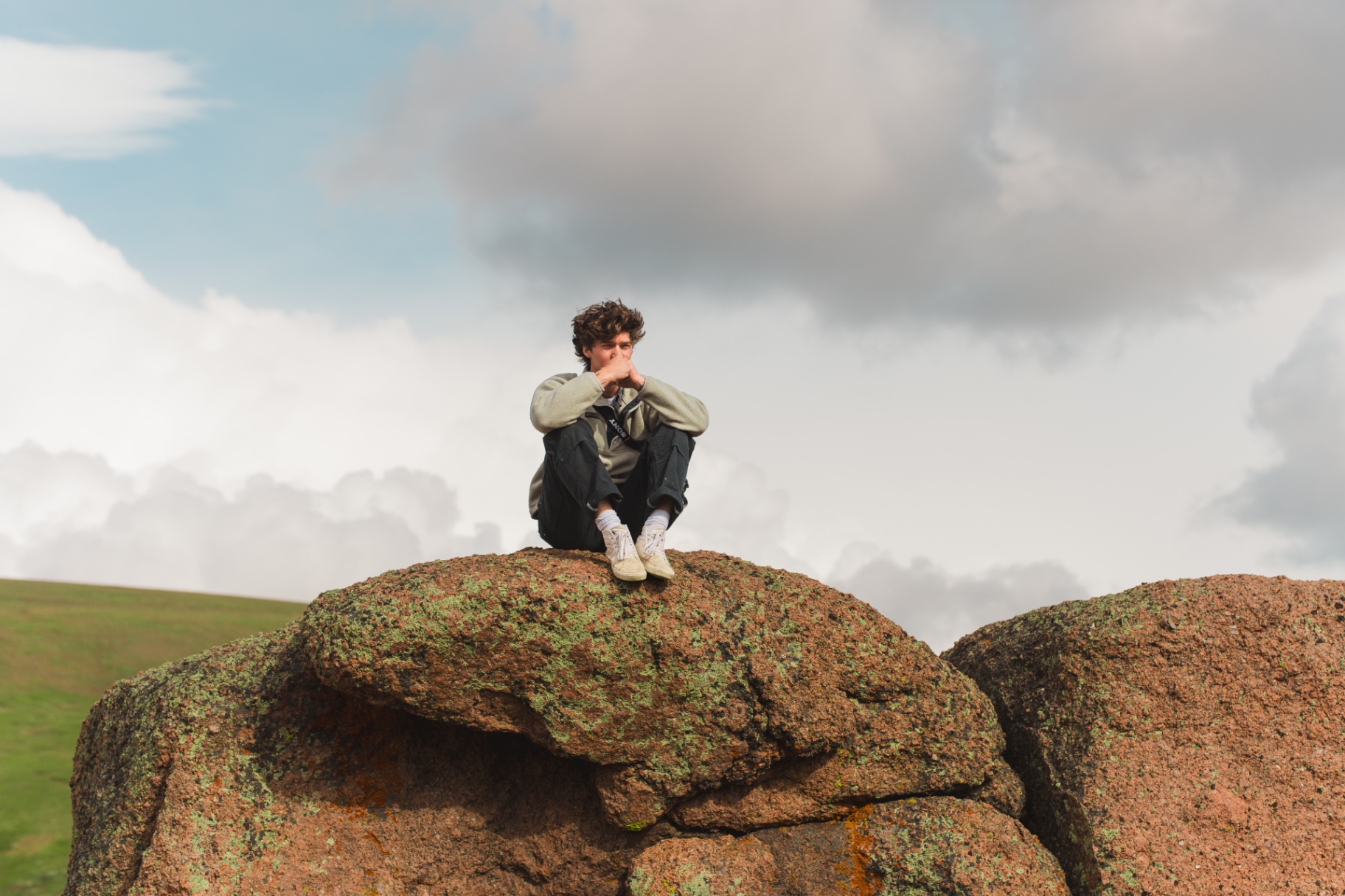 Young man seated atop a lichen-covered boulder beneath heavy clouds