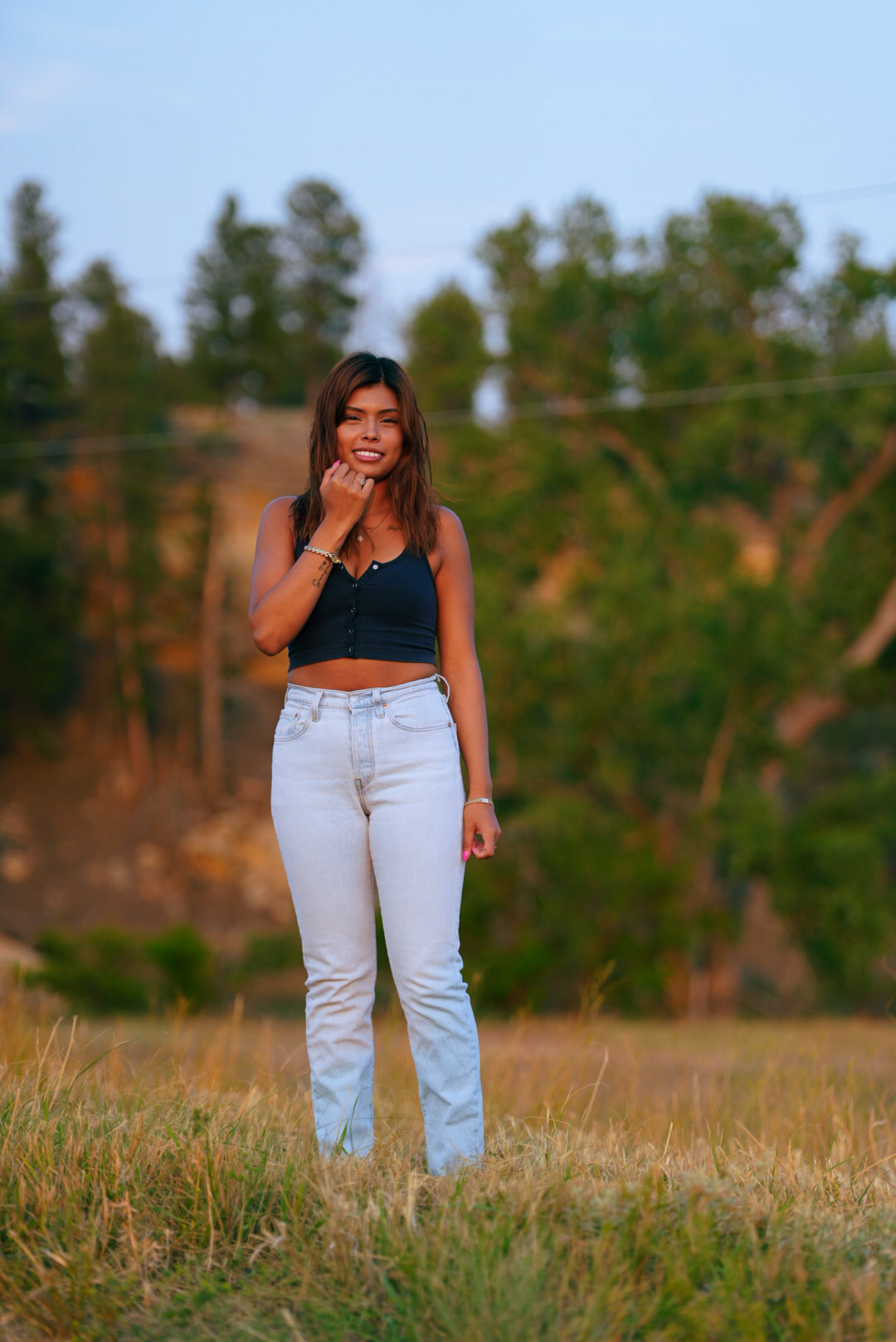 Woman standing in a dry grass field at golden hour framed by pines