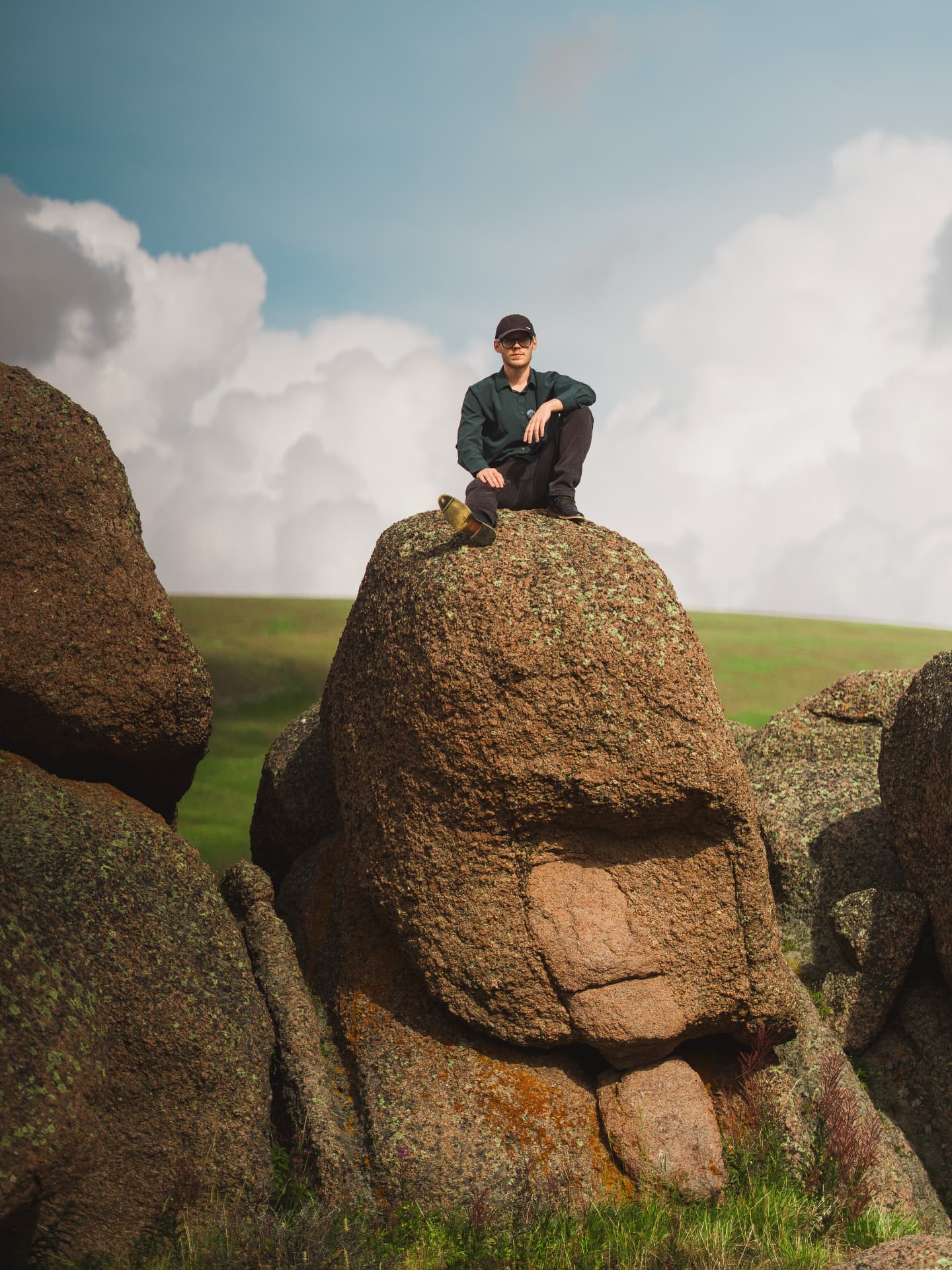 Man in a cap seated atop a lichen-covered granite boulder under storm clouds