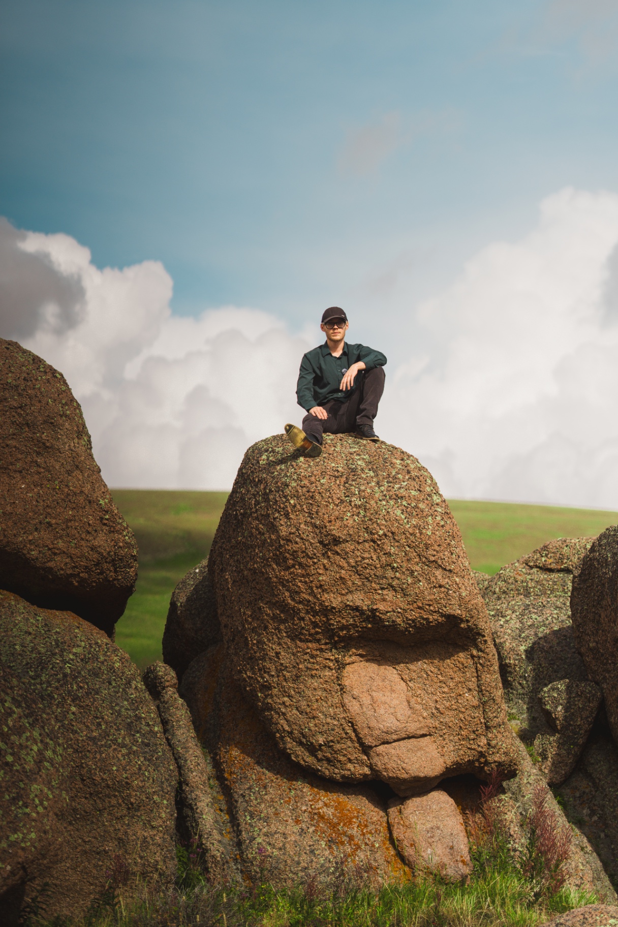 Man in a cap and sunglasses seated on a tall granite boulder under a blue sky