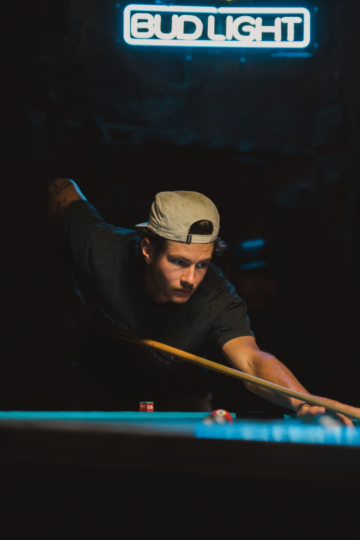 Man in a cap lining up a pool shot beneath a glowing Bud Light sign