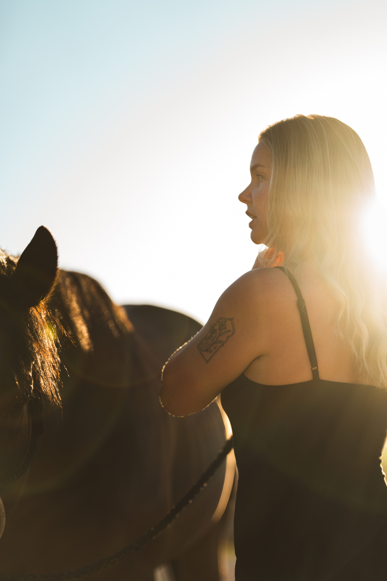 Blonde woman standing beside a horse backlit by bright sun