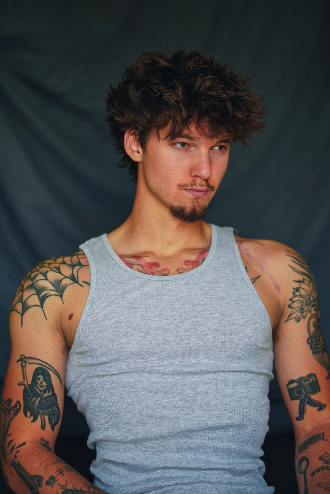 Studio portrait of a tattooed man in a grey tank top against a dark backdrop