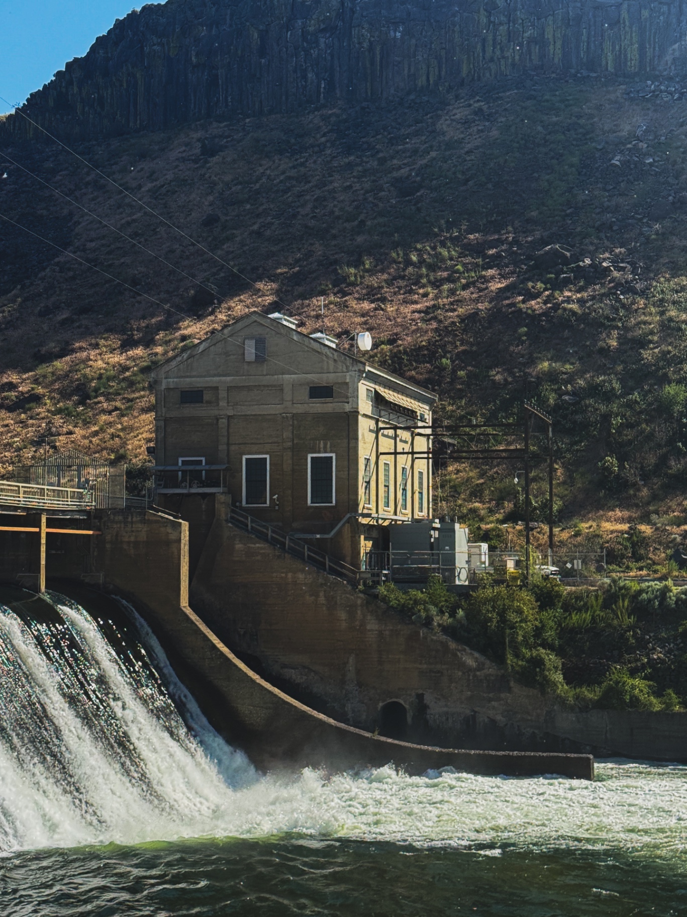 Historic powerhouse beside a spilling river dam under basalt cliffs