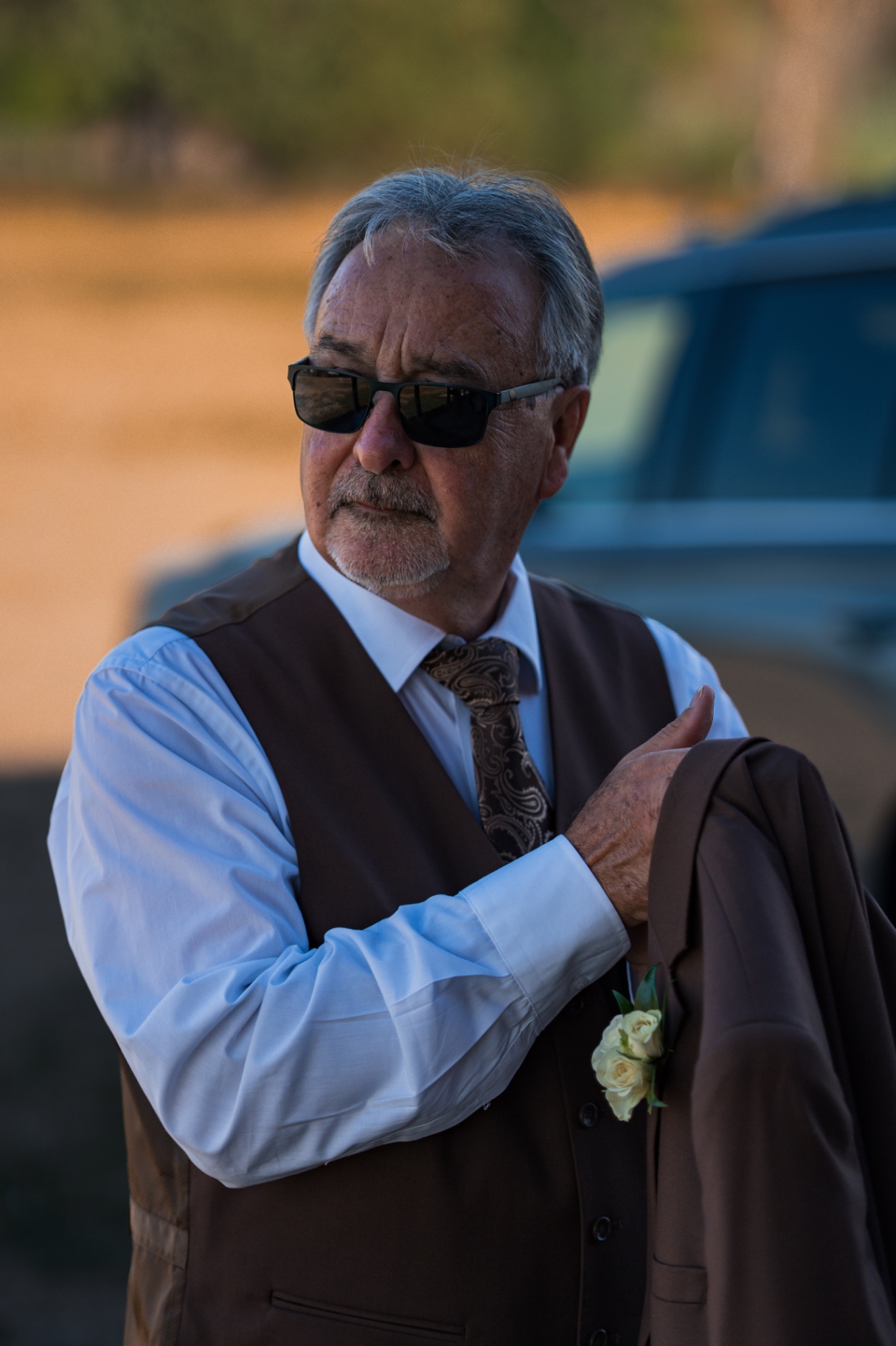 Older gentleman in a brown vest and sunglasses holding his jacket at sunset