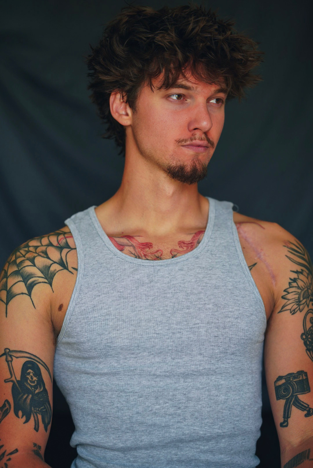 Tattooed man in a grey tank top looking off-camera against a dark backdrop