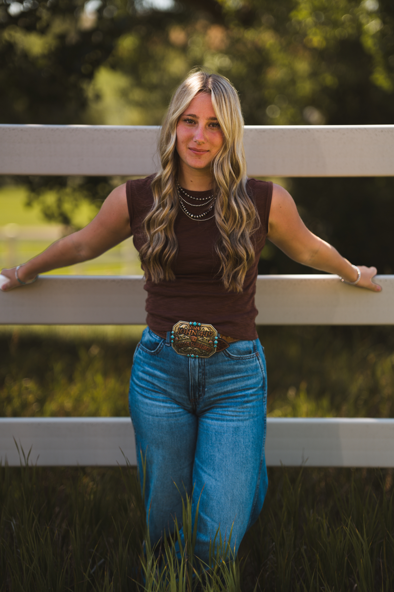 Blonde woman in a brown sleeveless top leaning against a white fence in warm sunlight
