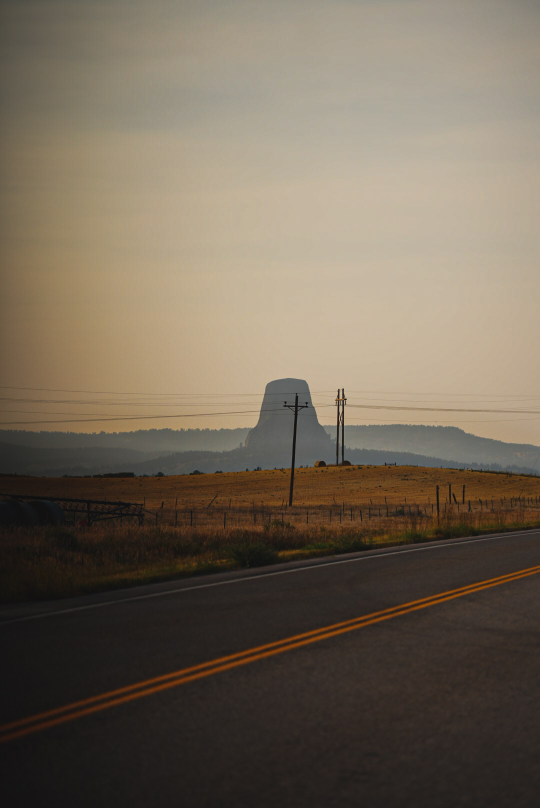 Devils Tower rising beyond power lines and a wheat field at dusk