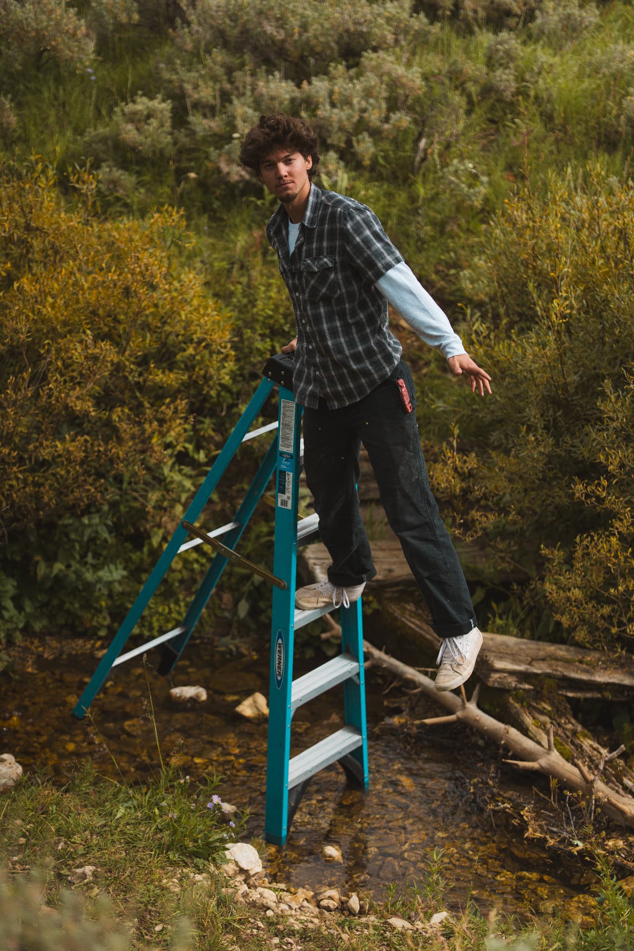 Man balancing on a teal step ladder set in a creek surrounded by brush