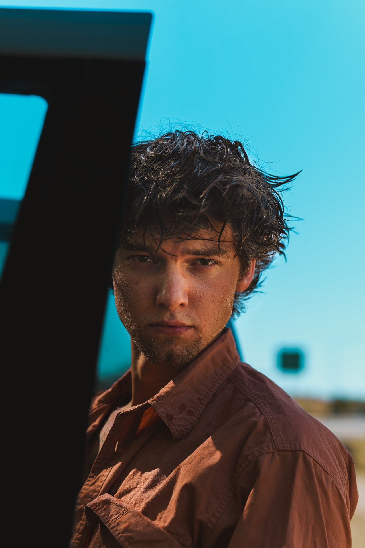 Young man with wind-tousled hair standing beside a vehicle against a clear blue sky
