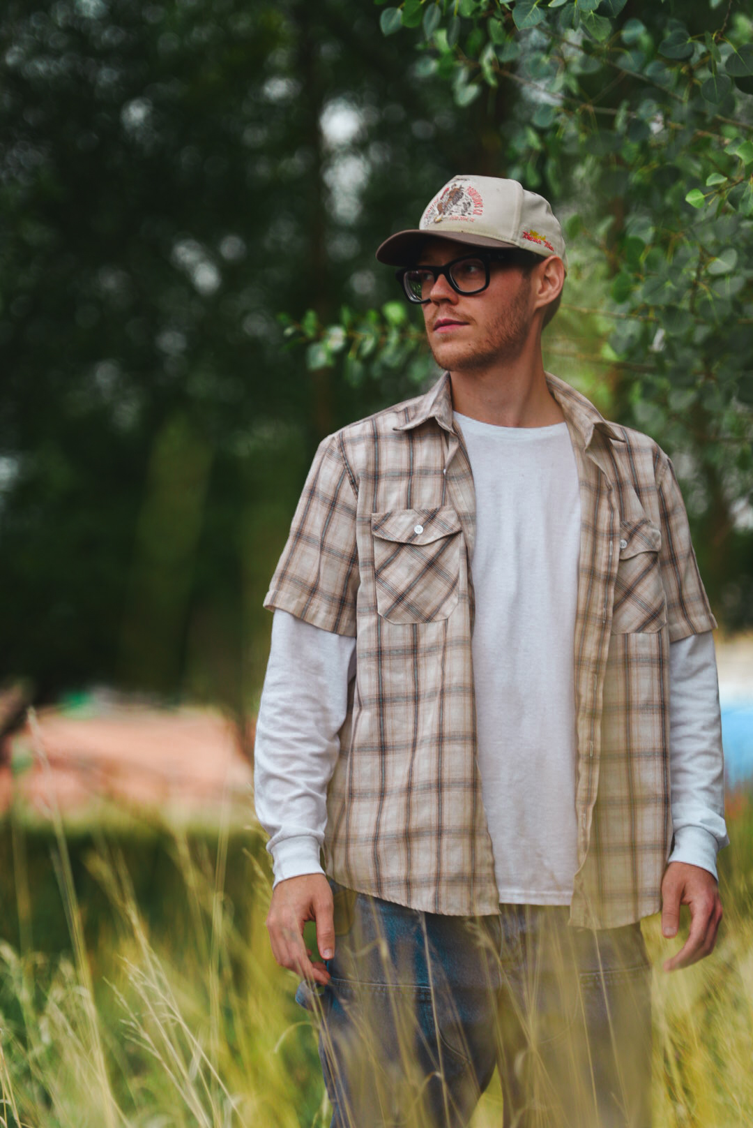 Man in a plaid shirt and cap looking off-camera framed by green leaves